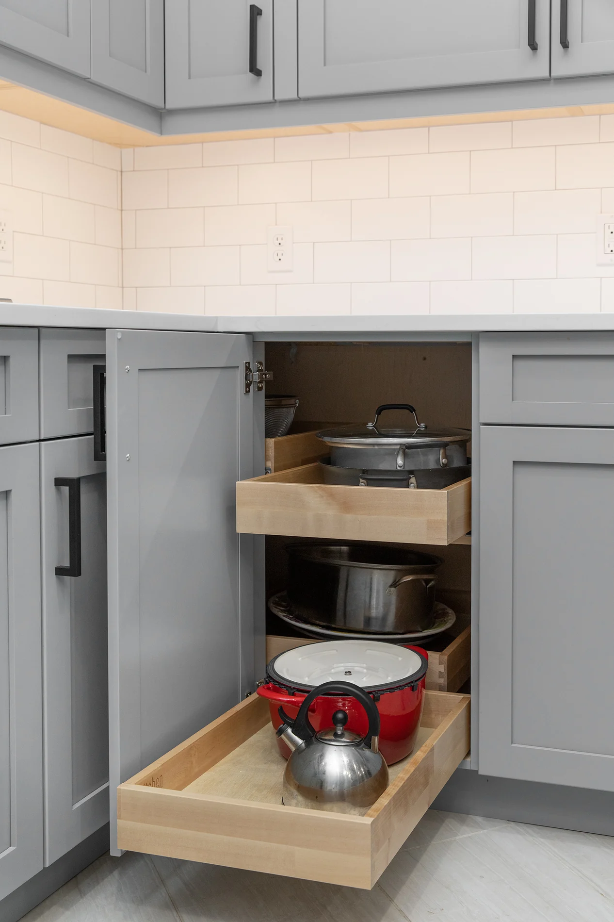 Corner base cabinet with two pull-out wood trays for pots and pans in a gray kitchen with white subway tile