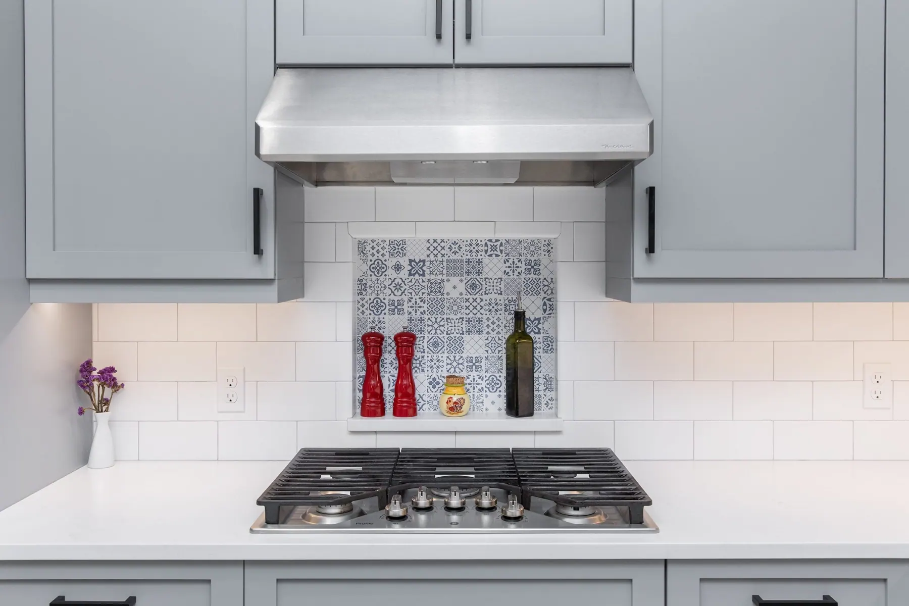 Cooktop under a stainless hood with a patterned blue and white tile niche in a gray shaker kitchen