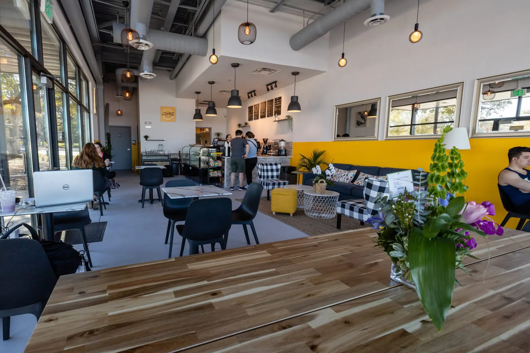 Cafe dining area with a wood table, black chairs, pendant lights, exposed ducts and a yellow accent wall