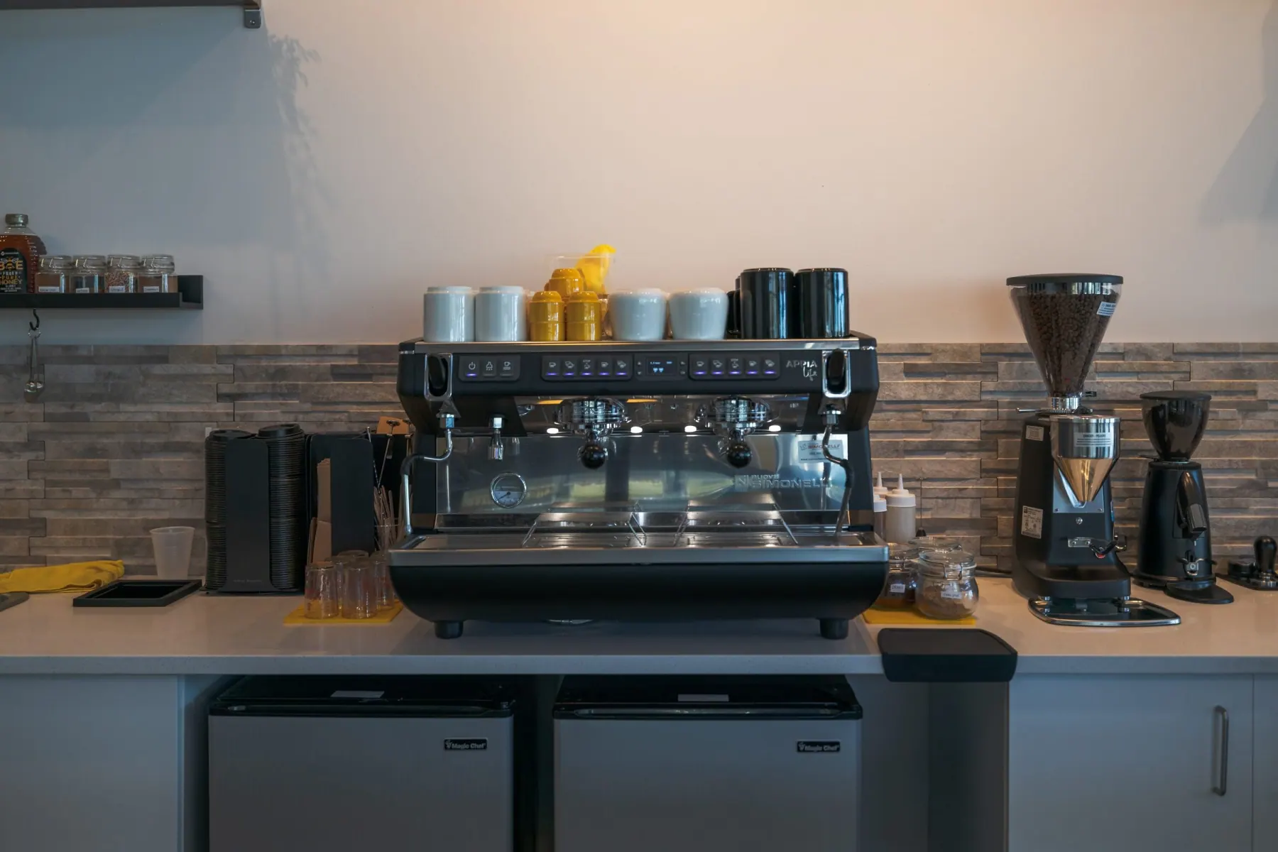 Espresso machine and grinders on a cafe counter with a stacked stone backsplash and two under-counter fridges