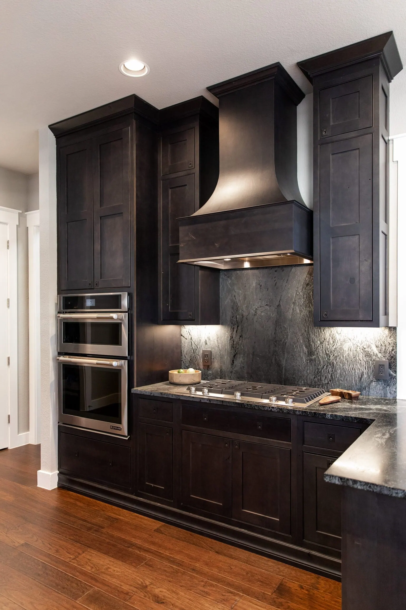 Dark-stained kitchen with a tapered wood hood, stacked wall ovens and hardwood floors, Westlake, Austin TX