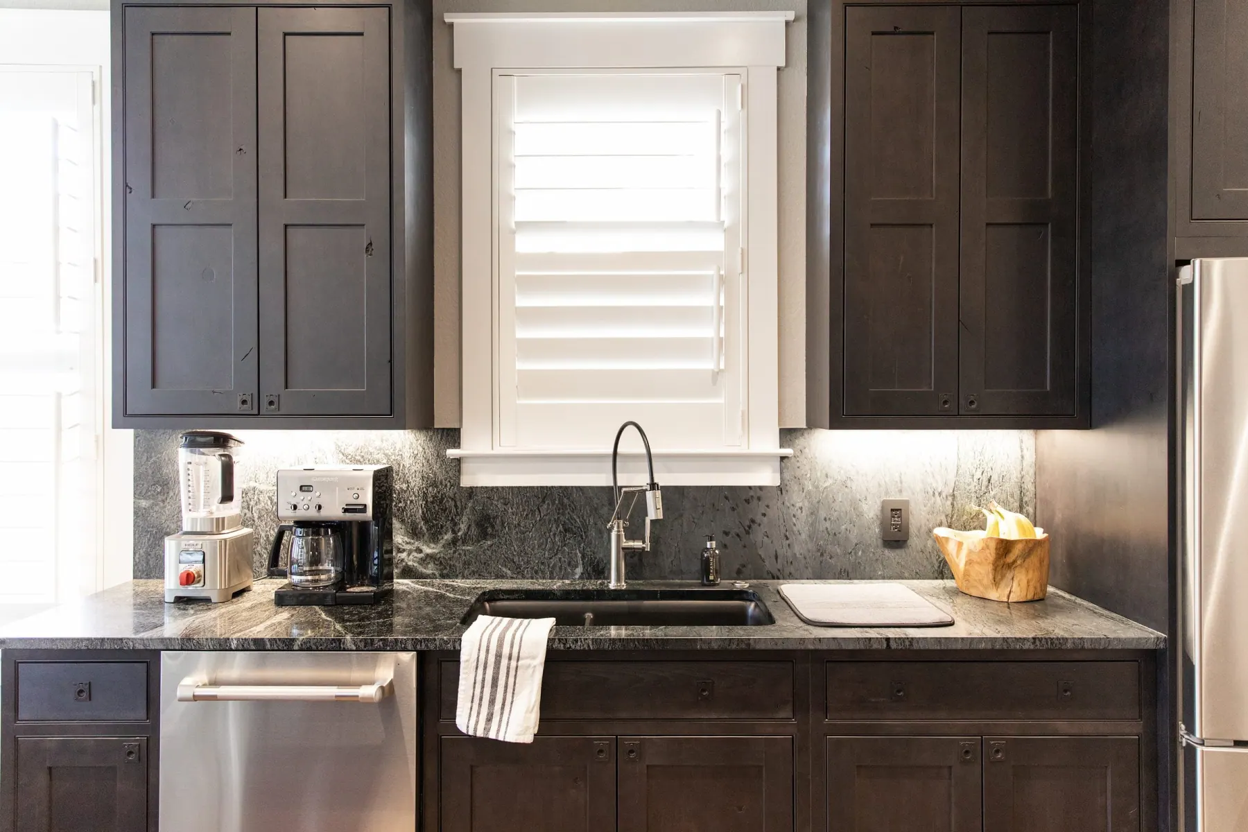 Kitchen sink wall with dark cabinets, white window shutters and a dark stone counter, Westlake, Austin TX